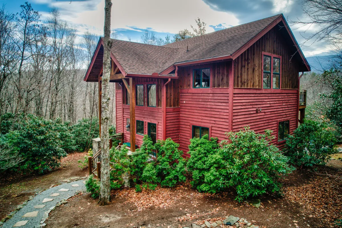 The red wooden exterior of Muir Lodge sits nestled among abundant greenery, with a stone pathway leading toward the entrance. The tranquil, cozy setting evokes the feeling of a mountain retreat surrounded by wilderness, inviting guests to begin their adventures right from the doorstep.