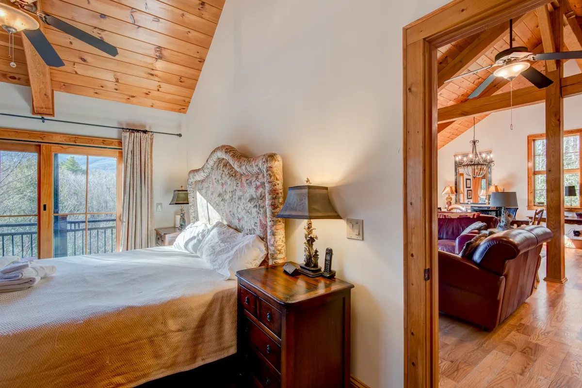 A bedroom interior showing a decorative headboard bed, nightstand with a lamp, and large windows framing a stunning natural landscape. The space feels airy and peaceful, ideal for relaxation, morning coffee, or enjoying a quiet evening.