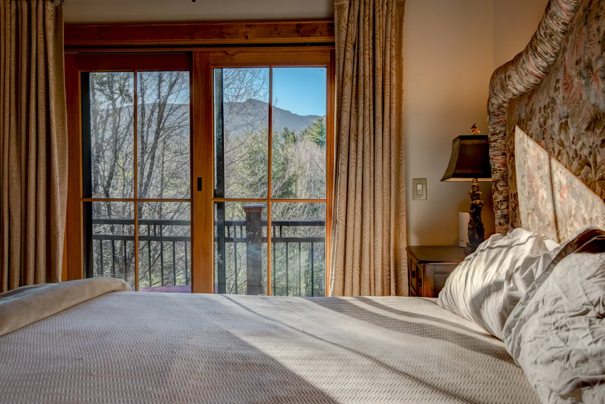 A serene downstairs bedroom featuring a comfortable-looking bed with soft linens, large window offering a view, and wooden furniture, creating an inviting, restful space. The window highlights direct access to the outdoors, reinforcing the seamless indoor-outdoor flow described in the section.