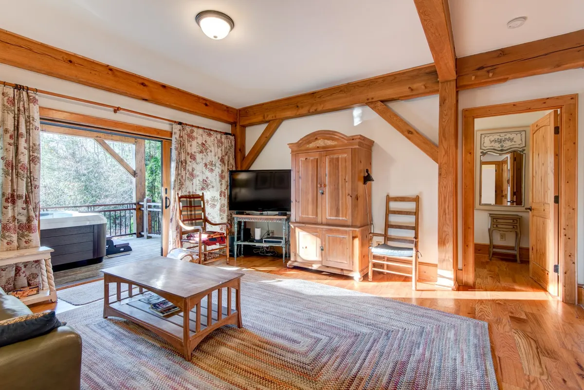 This image features a warm, inviting den area with comfortable seating, wooden beams, a large window letting in natural light, and an entertainment center. The space is clearly designed for relaxation and family gatherings, suggesting a perfect setting for movie nights, reading, or simply unwinding together.