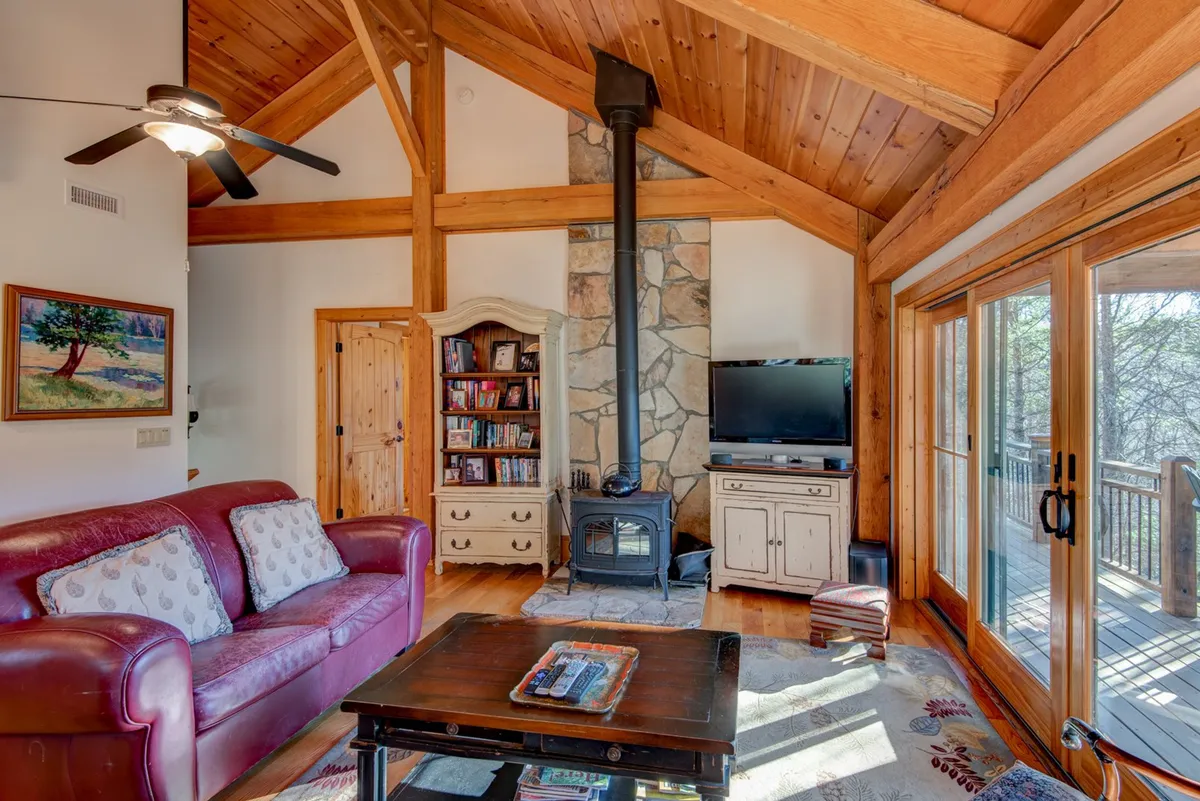A spacious great room featuring a red leather sofa, a stone fireplace ideal for cozy evenings, exposed wooden beams overhead, and large windows that flood the space with natural light and mountain views. The layout encourages group gatherings and fireside storytelling.