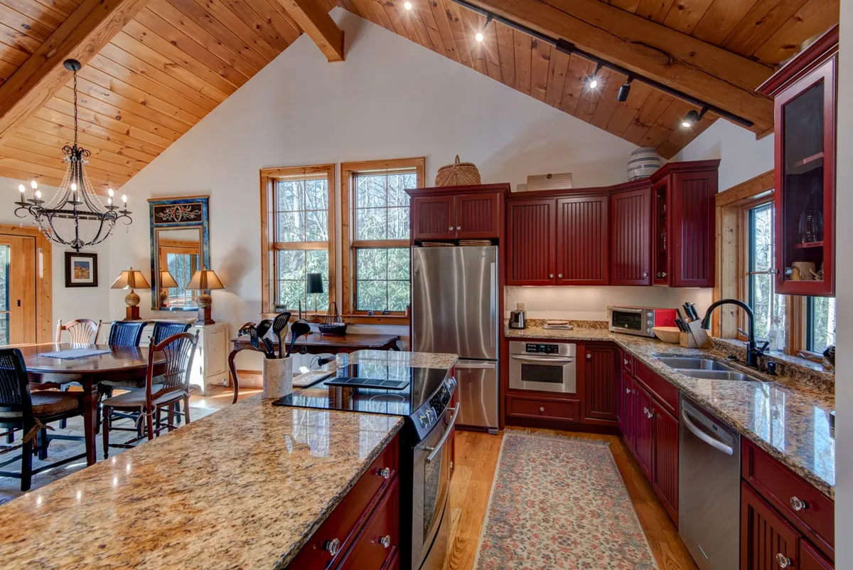 An open-plan space showcasing a granite kitchen island, sleek stainless steel appliances, and an adjacent wooden dining table set beneath rustic beams, with large windows framing mountain views. The kitchen and dining area are seamlessly integrated, perfect for shared meals and celebrations.