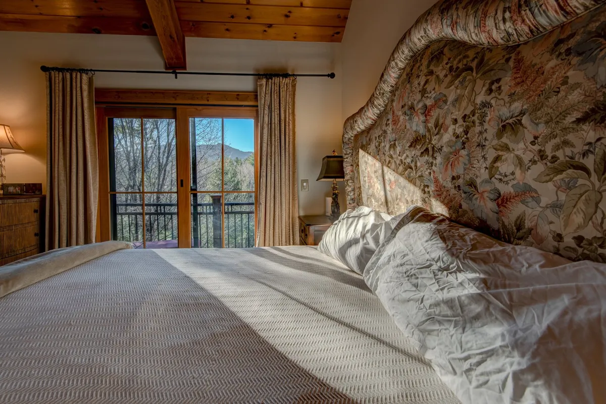 This interior shot features a master bedroom with a large floral headboard, exposed wooden beams and ceiling, balcony doors offering a direct mountain view, and soft, inviting bedding. The room is filled with natural light and offers a clear sense of privacy and tranquility, with the deck visible through the doors.