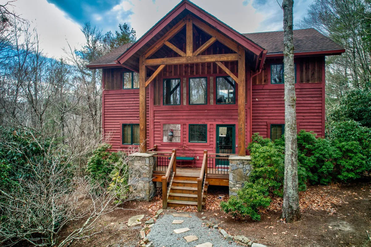 The hero image showcases Muir Lodge's stunning wooden architecture, featuring soaring posts and beams, large windows that invite natural light, and lush mountain greenery surrounding the home. A charming stone pathway leads up to the spacious porch, offering the perfect spot for relaxing, sipping morning coffee, or taking in breathtaking views of Grandfather Mountain. The scene radiates a warm, inviting atmosphere ideal for outdoor gatherings and unwinding in nature.