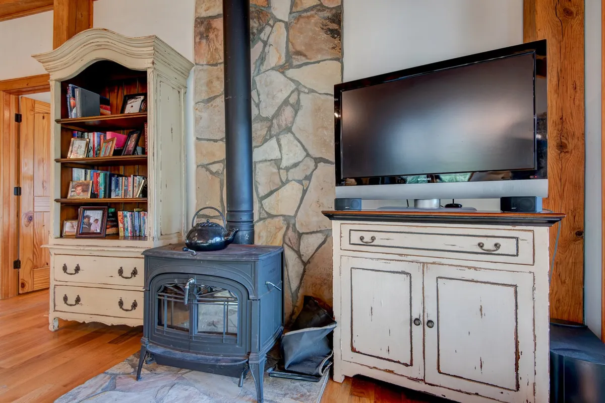 A cozy living area centers around a cast iron wood stove, with a bookshelf filled with books and a TV cabinet adding to the inviting atmosphere. The space is perfect for gathering with family after cold-weather adventures—think warming up with cocoa or reading by the fire after a day on the snowy trails. The decor and mood emphasize comfort and relaxation in a mountain retreat setting.
