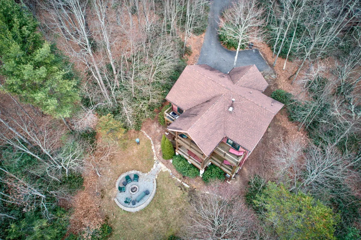 A cozy exterior shot featuring the house nestled among trees, with a focus on the circular fire pit and ample seating. The area is surrounded by nature, offering a tranquil setting ideal for gatherings and unwinding after outdoor adventures. The landscape’s gentle slope and openness reinforce the property's pet-friendly appeal.