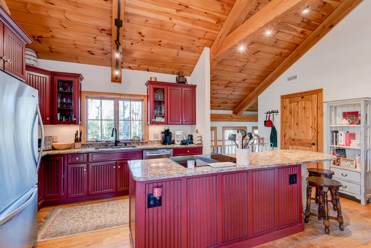 A welcoming kitchen scene highlighting a central island with granite countertops, red cabinetry, and large windows that let in natural light. The area is set up for cooking, casual dining, and entertaining guests.