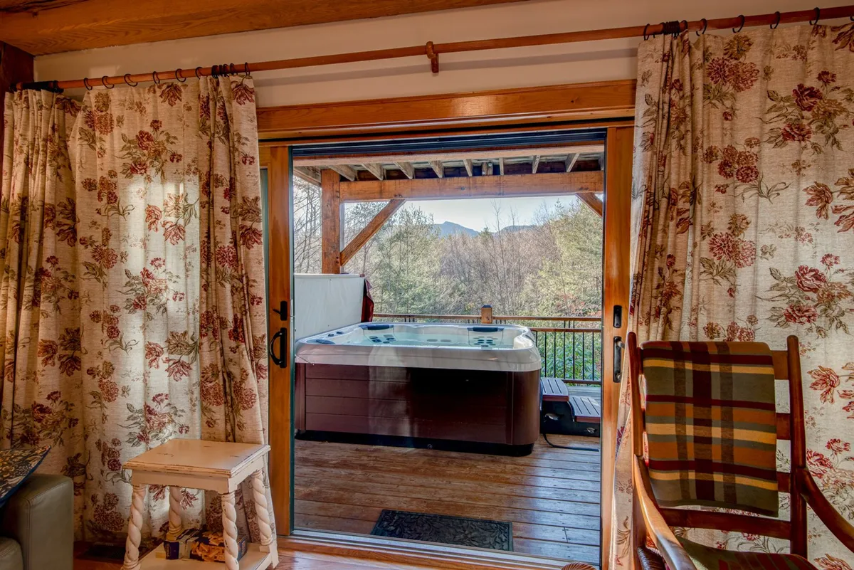 A tranquil hot tub scene set on a covered wooden deck, with curtains for privacy, a rocking chair, and views of the surrounding mountains. The inviting Jacuzzi is the focal point, surrounded by touches that hint at both relaxation and a connection to the outdoors.