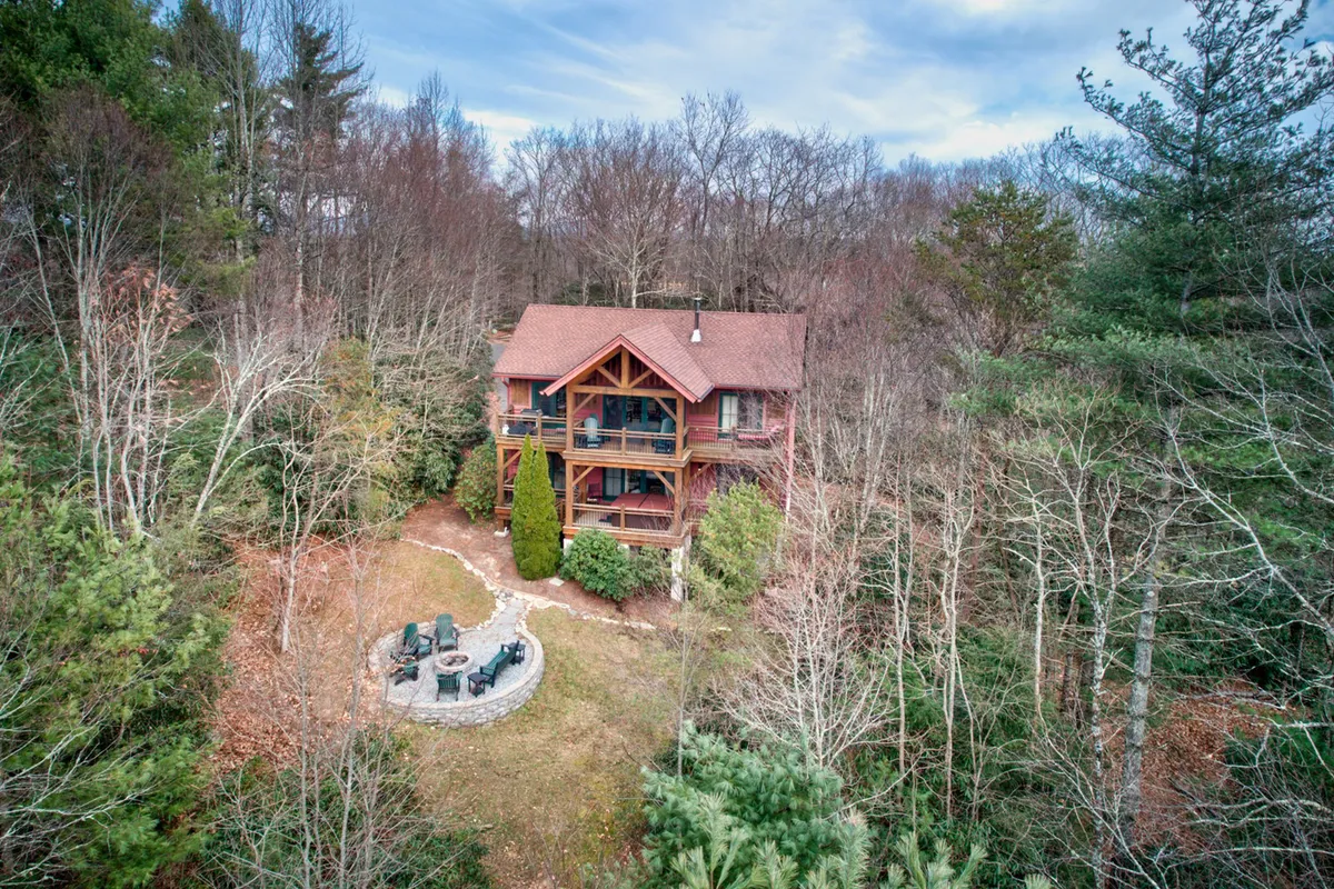 A wide exterior view of the house is surrounded by tall trees, showcasing both the structure and its natural, open setting. The fire pit and outdoor seating area are visible in the foreground, giving a sense of space for relaxing after a day of adventure. The lot appears nearly flat, making it ideal for pets to roam, and the setting sun creates a warm, inviting mood.