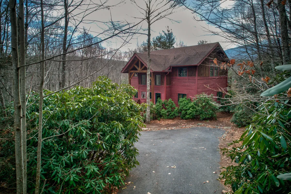 This image shows the red wooden exterior of Muir Lodge surrounded by lush green shrubbery, with a gently sloping, paved driveway leading directly to the front door. The home's inviting porch and scenic natural setting hint at both comfort and adventure, with easy access for unloading gear or welcoming pets after a day outside.