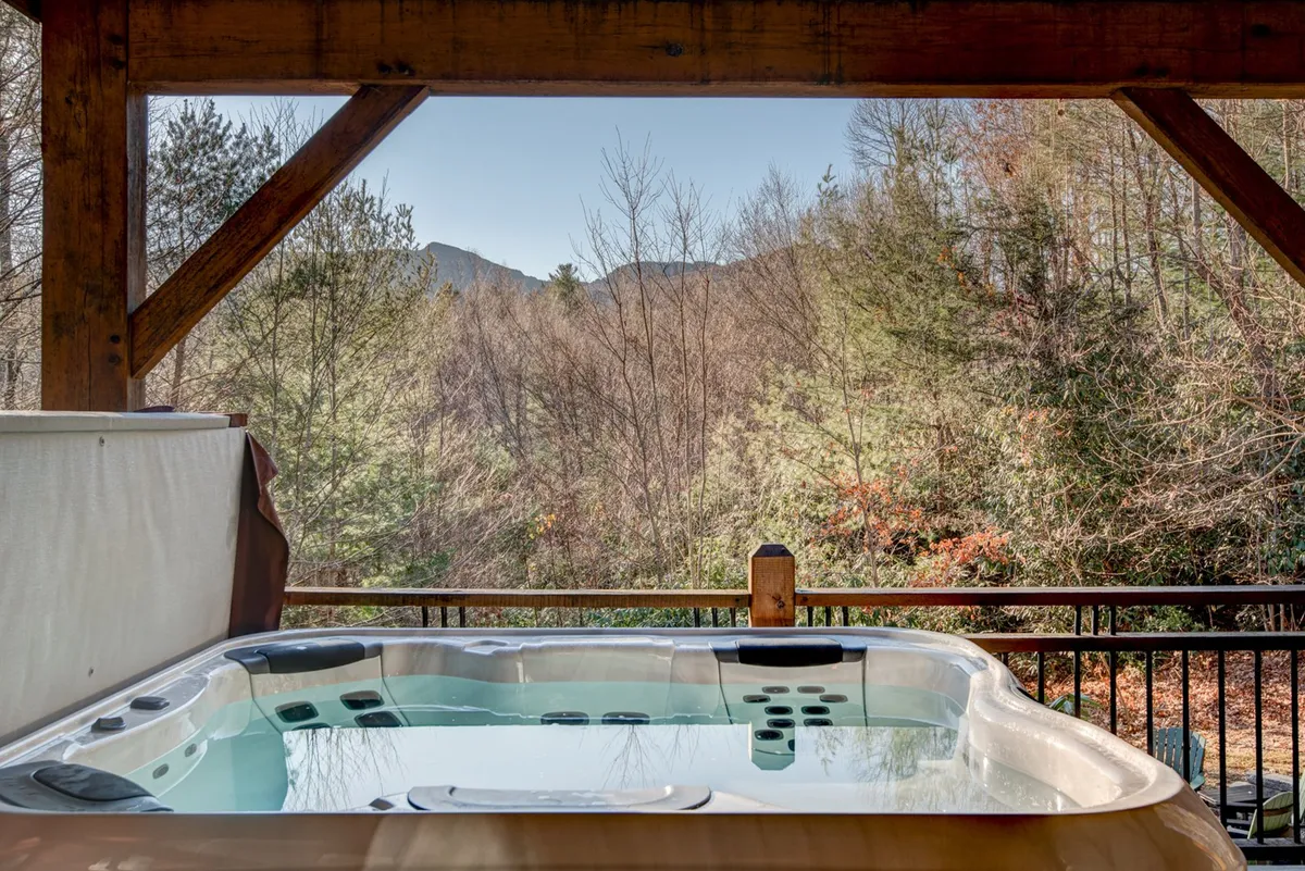 A tranquil outdoor hot tub set on a wooden deck, framed by lush trees and offering sweeping views of the mountains beyond. The scene evokes a sense of privacy and luxury, as the bubbling hot tub sits at the edge of nature, inviting guests to unwind while soaking in the panoramic vistas.