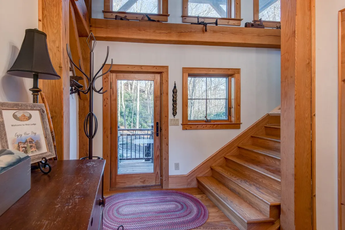 A welcoming entryway featuring a sturdy wooden staircase, front door, and warm decorative accents on a hallway table. Sunlight streams through nearby windows, hinting at the connection between indoors and the surrounding mountain setting. The scene exudes an inviting, transitional feel—perfect for guests arriving at the lodge after an outdoor adventure in any season.